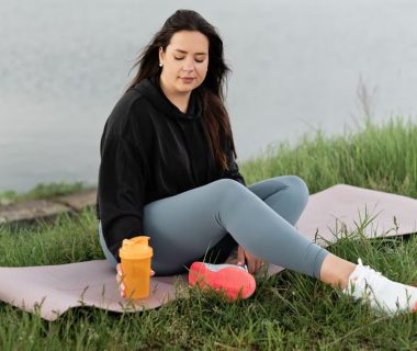 Woman enjoying a peaceful moment by the lake with a yoga mat and water bottle, promoting wellness.
